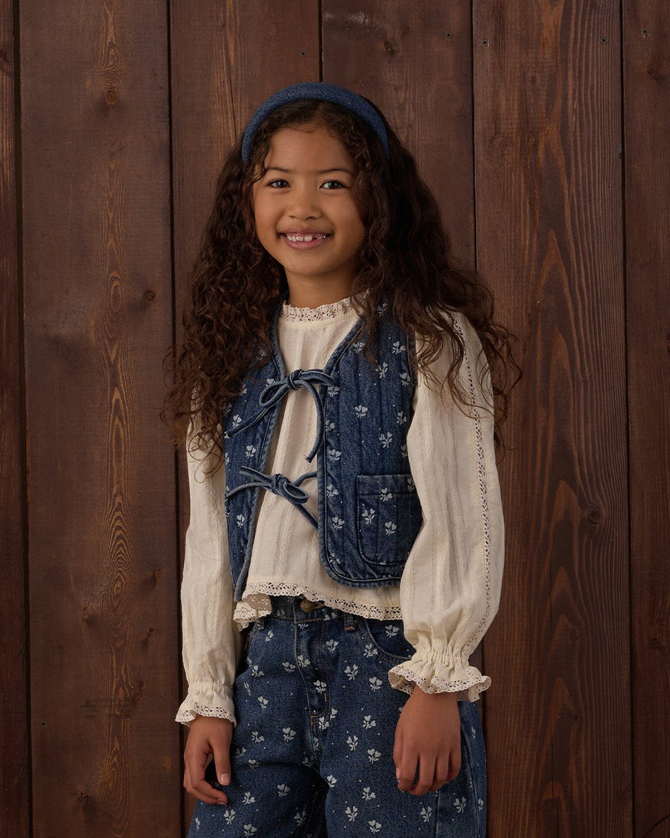 A young girl stands in front of a rustic dark wood plank wall wearing a vintage-style outfit consisting of a cream ruffled blouse, a blue floral vest, matching blue floral pants, and black shoes. The scene is styled with a wooden chair leg, pinecones, a lantern, and stacked logs on the floor.