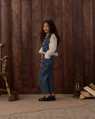 A young girl stands in profile against a dark brown wooden plank wall, wearing a blue floral print pinafore-style romper over a white long-sleeve blouse, with a blue headband and black shoes.