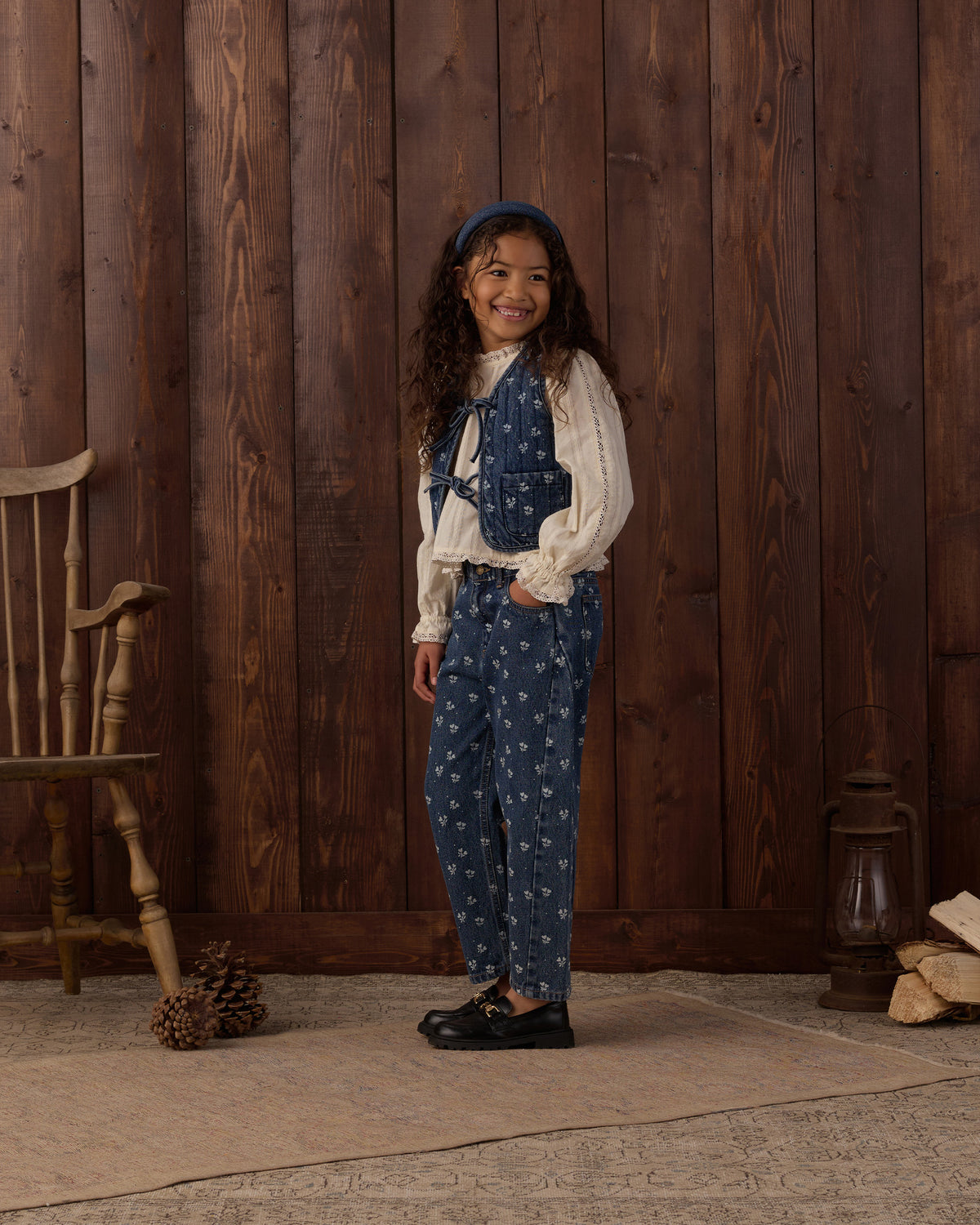A young girl stands in front of a rustic dark wood plank wall wearing a blue patterned vest and matching pants over a cream puff-sleeve blouse, with a blue headband and black shoes. A wooden chair, pinecones, and a lantern are visible in the scene.