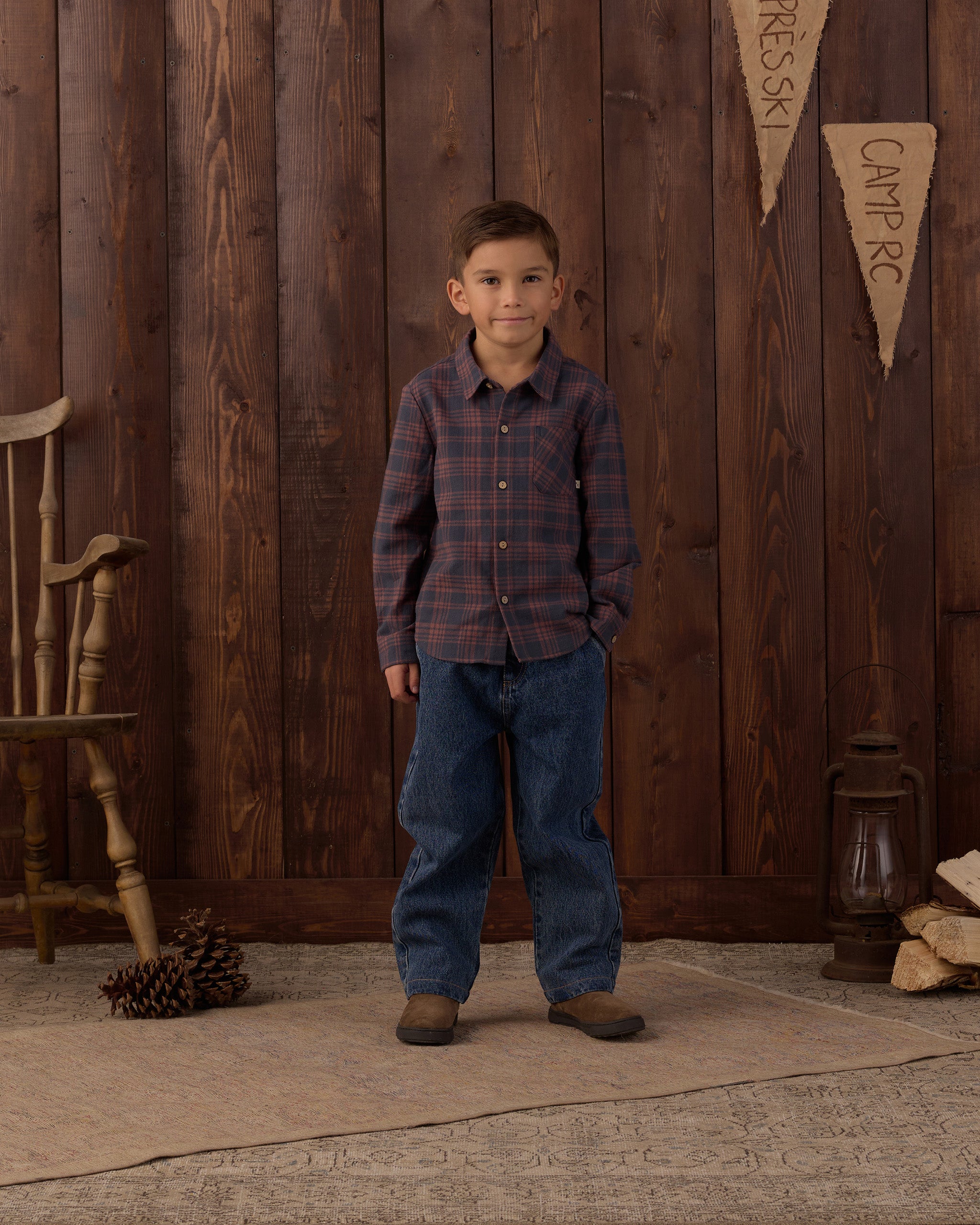 A young boy stands in front of a rustic dark wood plank wall wearing a long-sleeve plaid button-down shirt in muted blue and red tones, paired with blue jeans and brown boots. A wooden chair, pinecone, lantern, and stacked wood are visible in the scene.