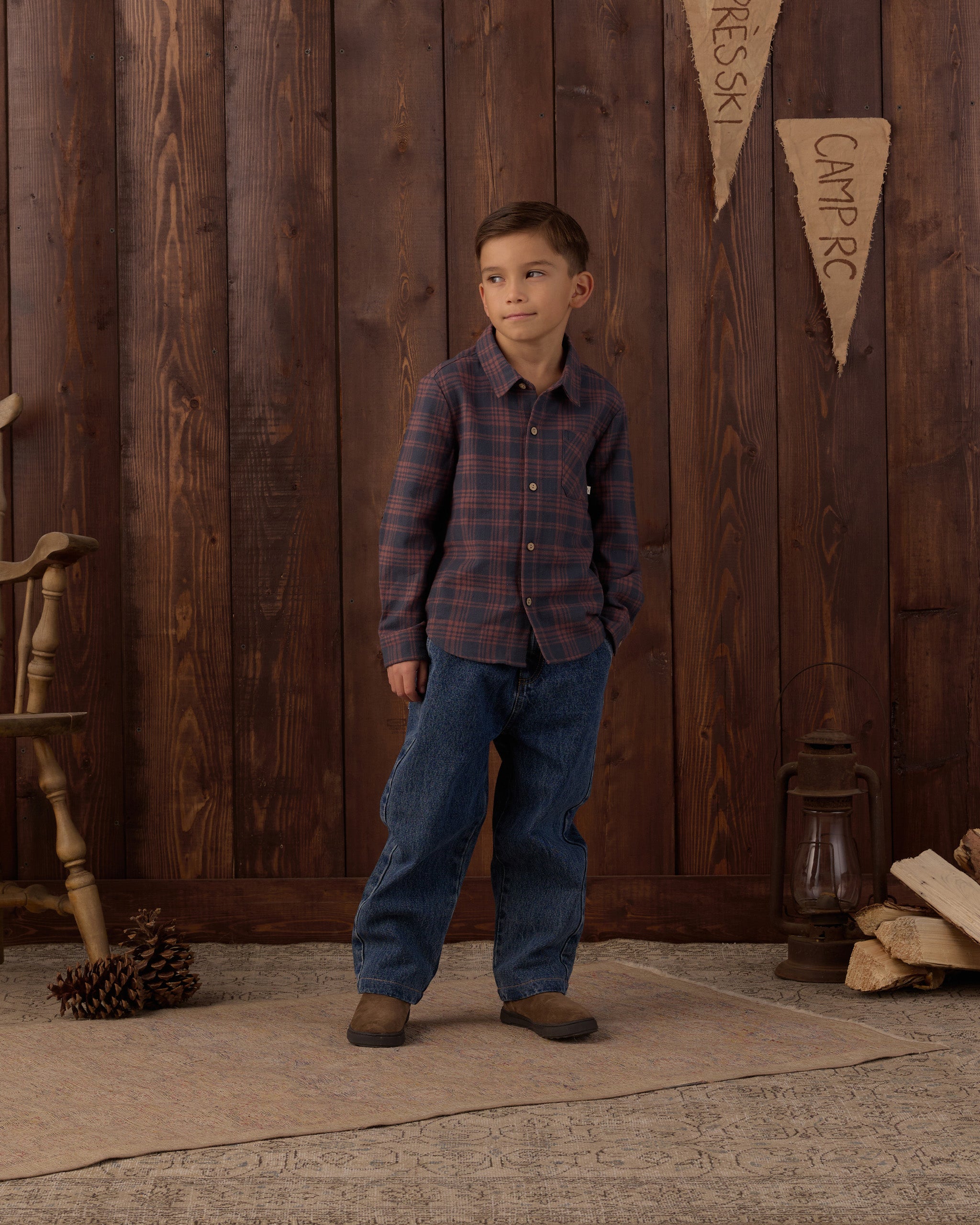 A young boy stands in front of a rustic dark wood plank wall wearing a long-sleeve plaid button-up shirt in dark red and navy, blue jeans, and brown boots. Decorative elements like a wooden chair, a lantern, pinecones, and a small pennant sign are visible on the floor and wall.