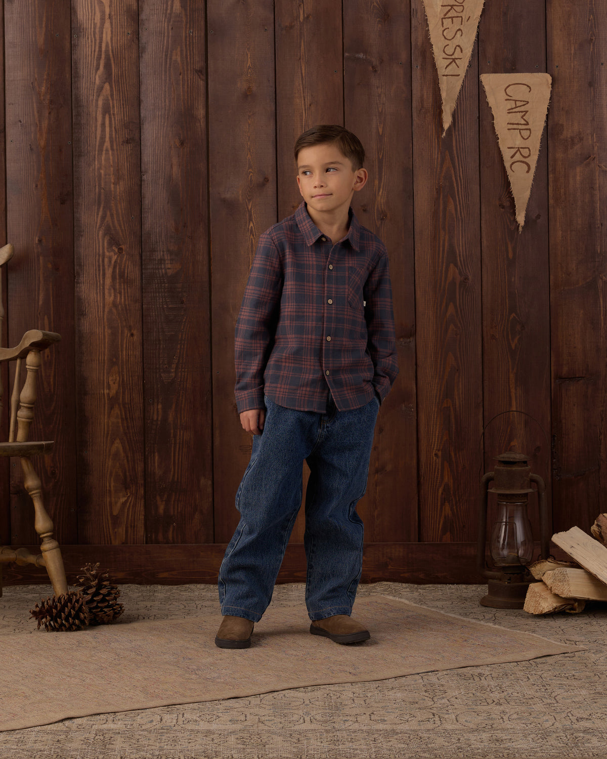 A young boy stands in front of a rustic dark wood plank wall wearing a long-sleeve plaid button-up shirt in dark red and navy, blue jeans, and brown boots. Decorative elements like a wooden chair, a lantern, pinecones, and a small pennant sign are visible on the floor and wall.