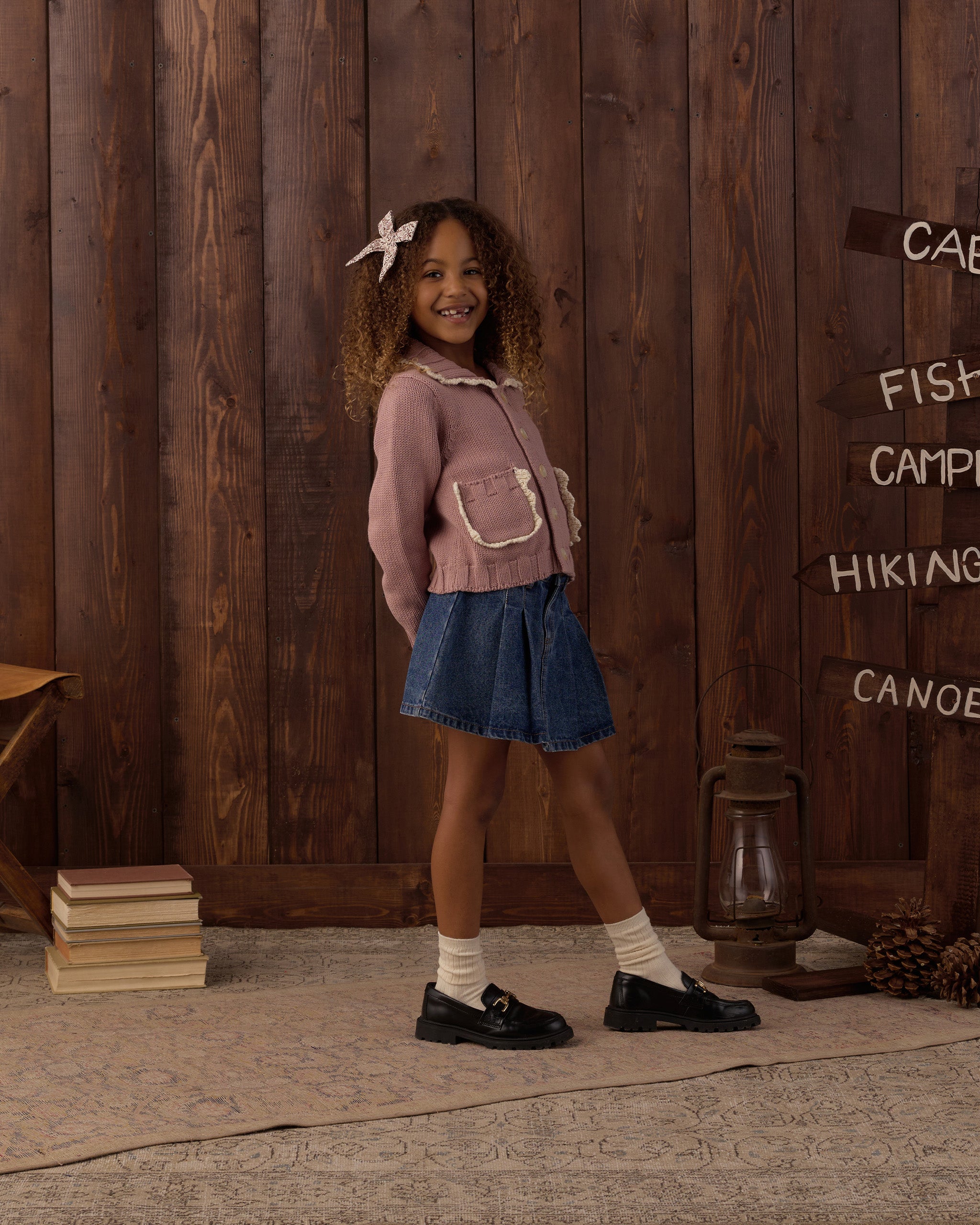 A young girl stands in front of a rustic dark wood plank wall wearing a dusty pink knit cardigan with white trim, a blue denim skirt, white socks, and black loafers. The background features a vintage-style lantern, a stack of books, and directional signs pointing to outdoor activities.
