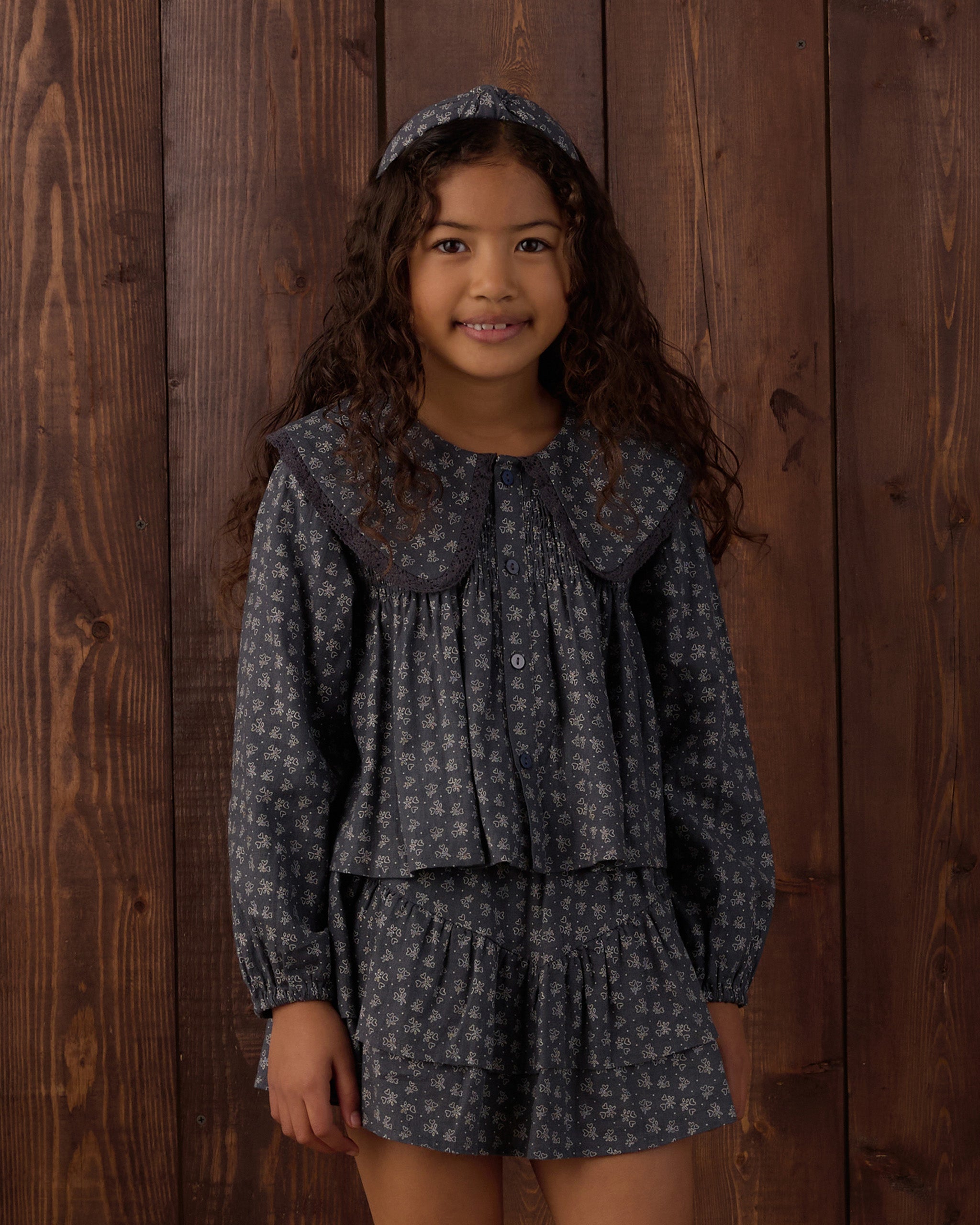 A young girl with long curly brown hair stands against a dark wood plank wall wearing a matching dark gray and white floral print outfit consisting of a long-sleeve button-front top with a large Peter Pan collar, a matching ruffled mini skirt, and a coordinating headband.