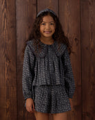 A young girl with long curly brown hair stands against a dark wood plank wall wearing a matching dark gray and white floral print outfit consisting of a long-sleeve button-front top with a large Peter Pan collar, a matching ruffled mini skirt, and a coordinating headband.