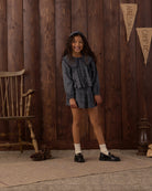 A young girl stands in front of a rustic dark wood plank wall wearing a matching long-sleeve ruffled top and mini skirt set in a dark blue-gray and white pattern. She is styled with white socks and black loafers, with a wooden chair, pinecones, and a lantern visible in the background.
