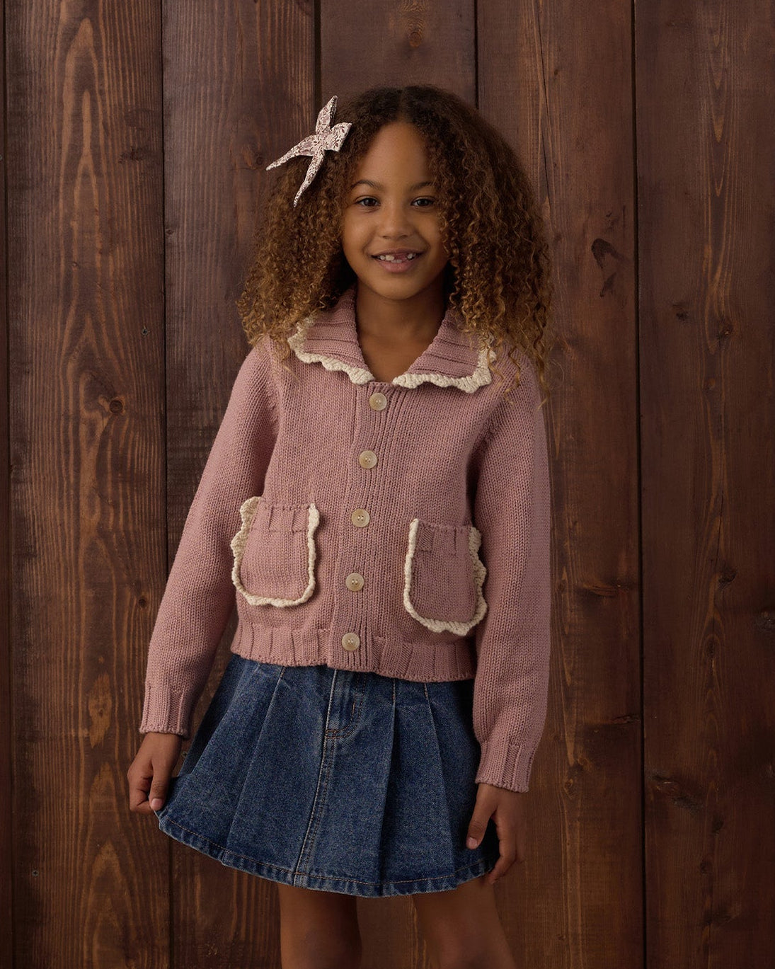 A young girl stands in front of a rustic dark wood plank wall wearing a dusty pink cardigan with a ruffled cream collar, a blue denim mini skirt, white socks, and black shoes.