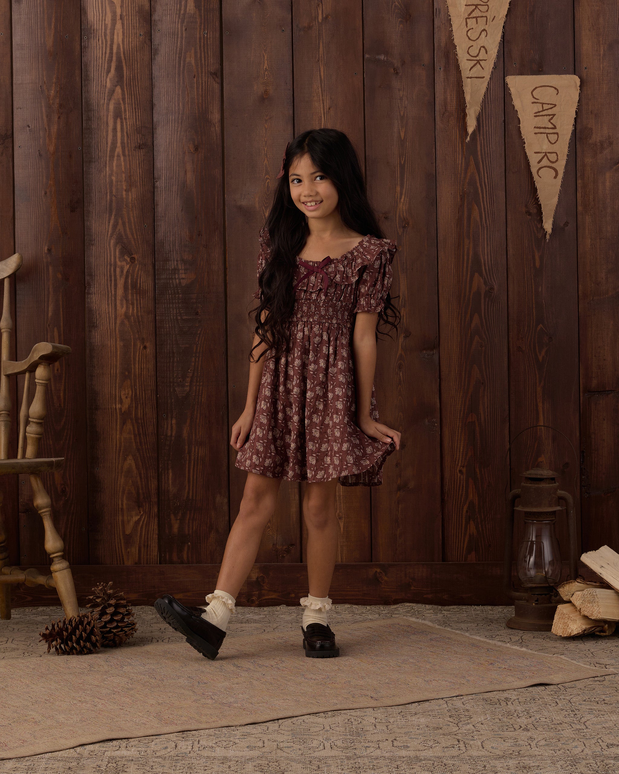 A young girl with long dark hair stands in a rustic room with dark wood paneling, wearing a short-sleeved smocked dress with a dark red and brown floral pattern. She is wearing white socks and black shoes, standing on a textured rug next to a wooden chair and a lantern.