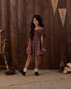 A young girl with long dark hair stands in a rustic room with dark wood paneling, wearing a short-sleeved smocked dress with a dark red and brown floral pattern. She is wearing white socks and black shoes, standing on a textured rug next to a wooden chair and a lantern.