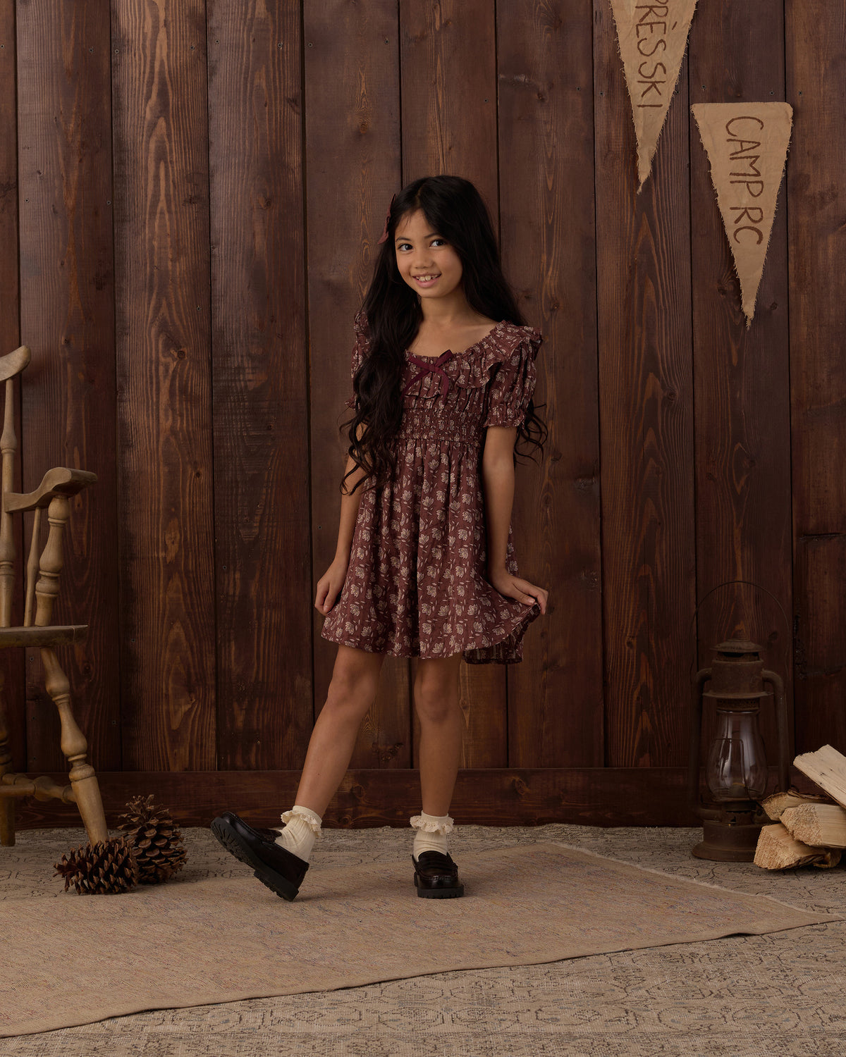 A young girl with long dark hair stands in a rustic room with dark wood paneling, wearing a short-sleeved smocked dress with a dark red and brown floral pattern. She is wearing white socks and black shoes, standing on a textured rug next to a wooden chair and a lantern.