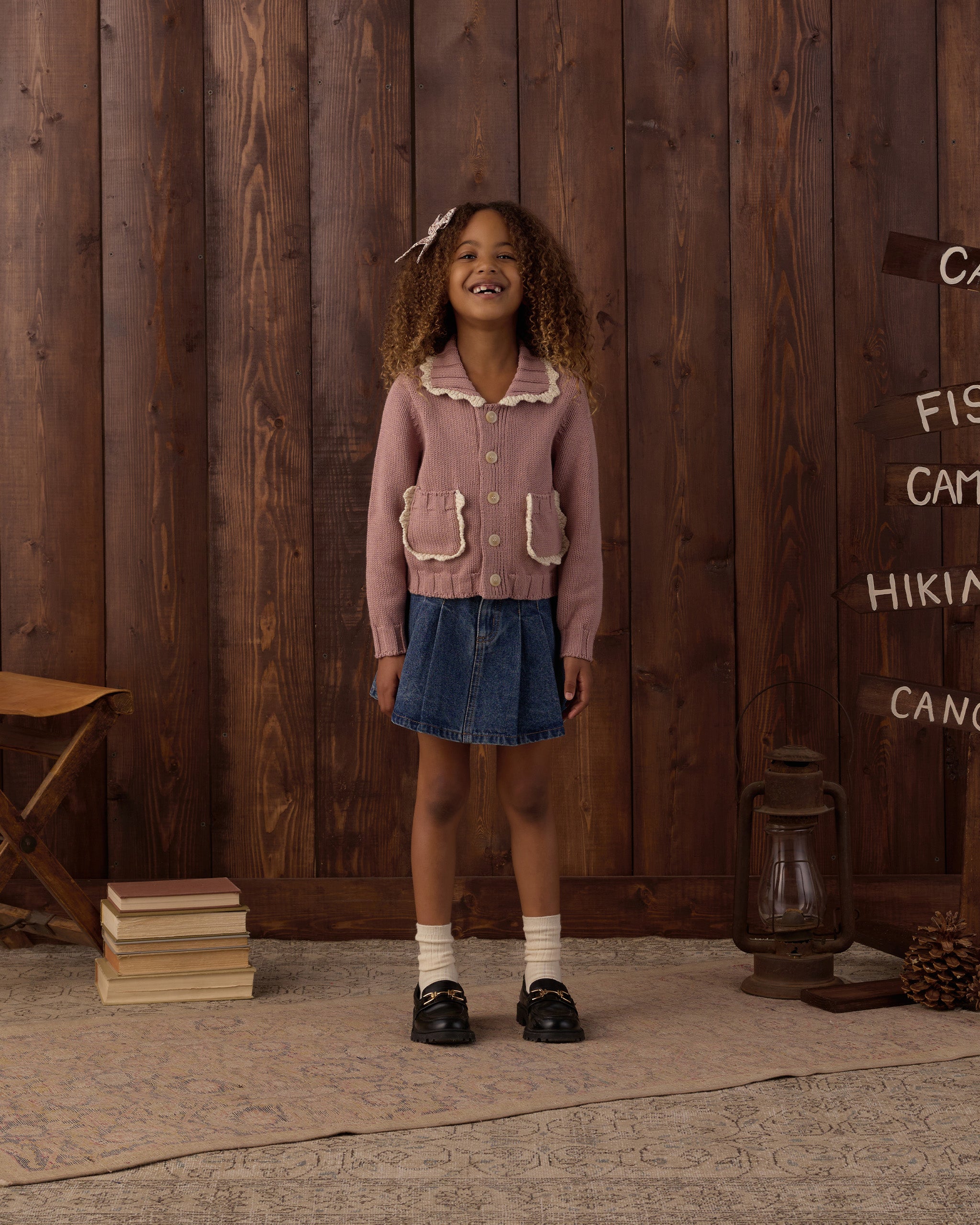A young girl stands in front of a rustic dark wood plank wall wearing a dusty pink knit cardigan with cream ruffle trim and front pockets, a blue denim skirt, white socks, and black shoes.