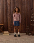 A young girl stands in front of a rustic dark wood plank wall wearing a dusty pink knit cardigan with cream ruffle trim and front pockets, a blue denim skirt, white socks, and black shoes.