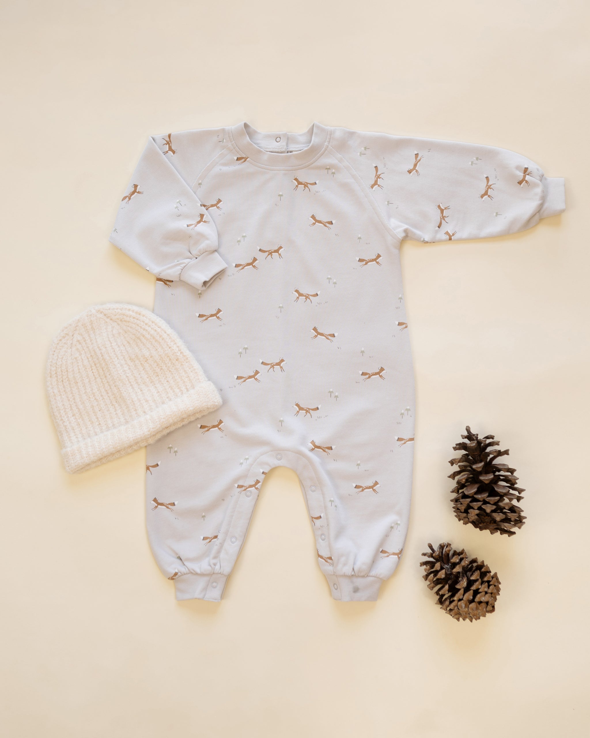 A baby long-sleeve footless onesie in white with a small brown animal print, laid flat on a cream background alongside a white knit beanie and two pinecones.