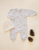 A baby long-sleeve footless onesie in white with a small brown animal print, laid flat on a cream background alongside a white knit beanie and two pinecones.