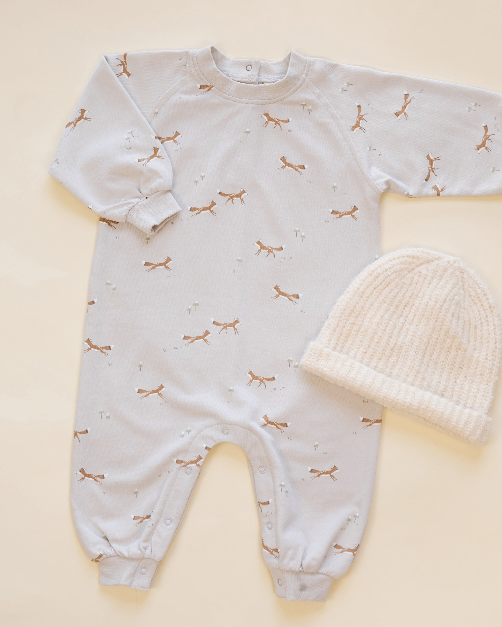 A baby's long-sleeve footless onesie in white with a delicate brown and gold butterfly print, shown next to a cream-colored knit baby bonnet on a light beige background.