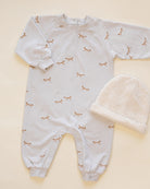 A baby's long-sleeve footless onesie in white with a delicate brown and gold butterfly print, shown next to a cream-colored knit baby bonnet on a light beige background.