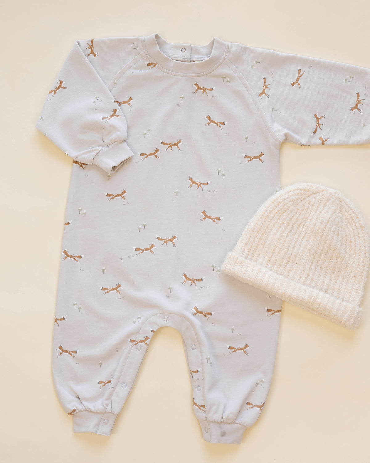 A baby's long-sleeve footless onesie in white with a delicate brown and gold butterfly print, shown next to a cream-colored knit baby bonnet on a light beige background.