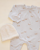 A baby long-sleeve footless romper in a very pale pink or off-white, printed with small brown foxes and tiny white stars, shown flat-laid next to a cream knit beanie on a warm cream background.