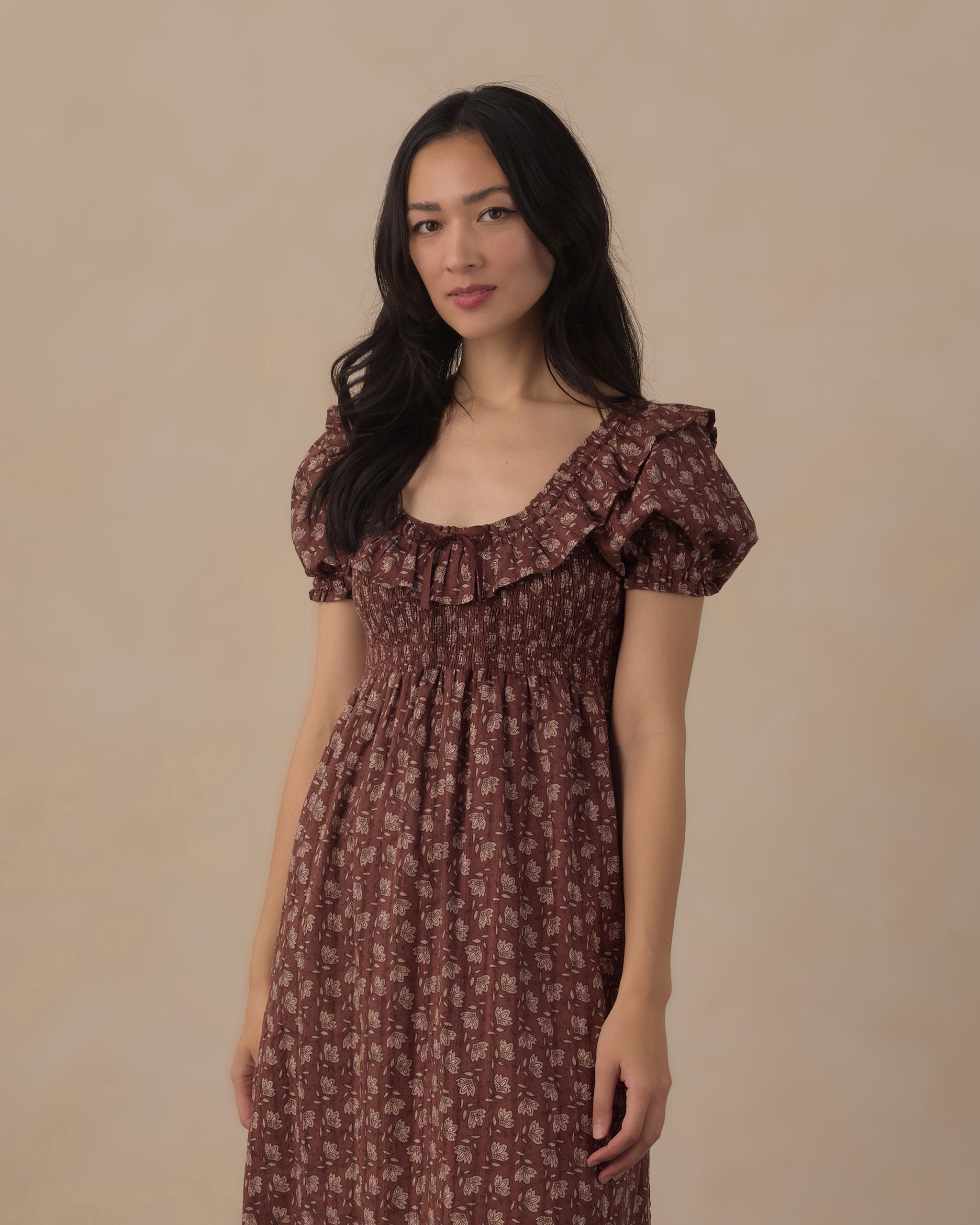 A woman wearing a brown and beige patterned midi dress with puff sleeves and a smocked bodice, standing against a solid beige background.