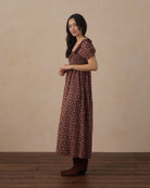 A woman modeling a brown short-sleeve maxi dress with a small floral pattern, standing against a neutral beige wall and wooden floor.