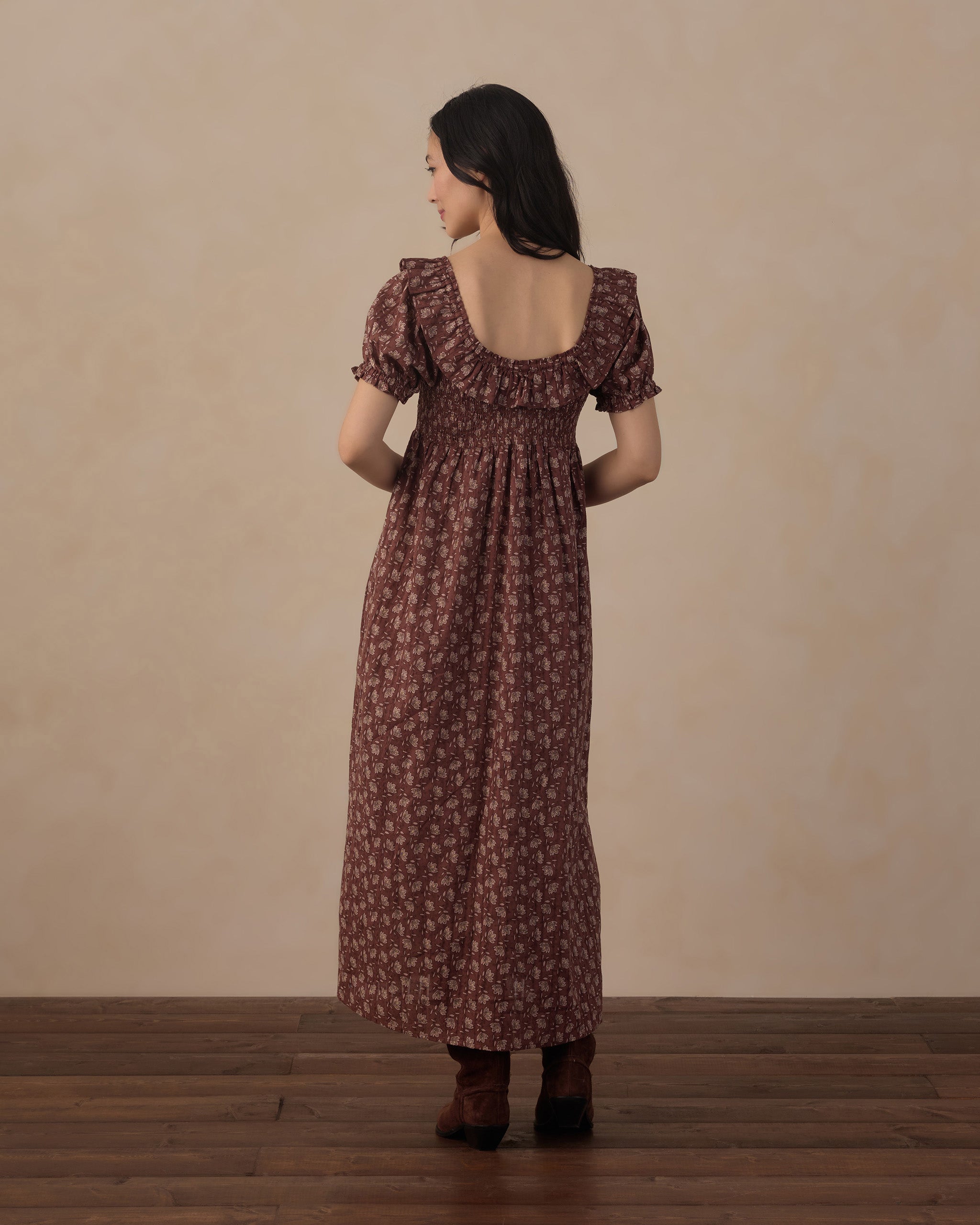 A woman stands with her back to the camera wearing a dark brown midi dress with a small floral pattern and puff sleeves, paired with dark brown boots, set against a plain beige wall and wooden floor.