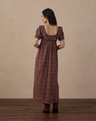 A woman stands with her back to the camera wearing a dark brown midi dress with a small floral pattern and puff sleeves, paired with dark brown boots, set against a plain beige wall and wooden floor.