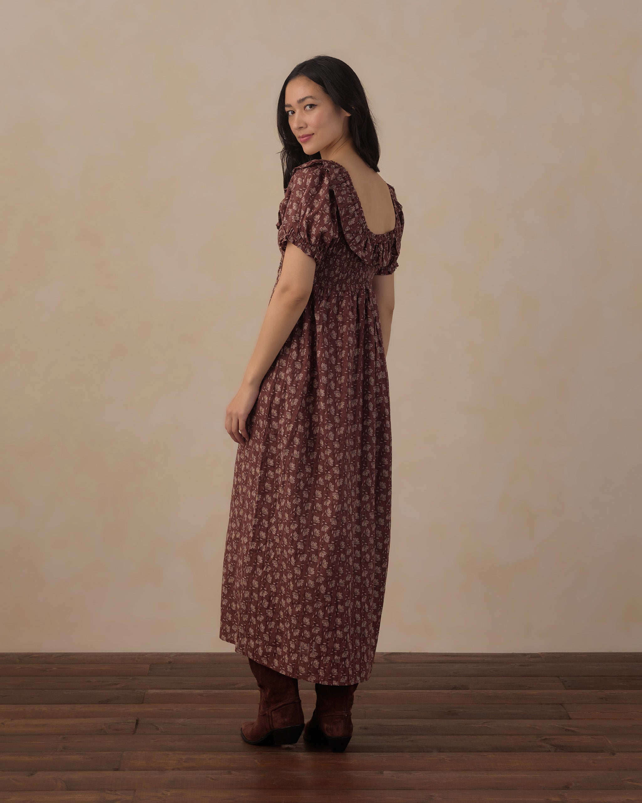 A woman wearing a brown and beige floral midi dress with puff sleeves and a low back, standing in profile against a neutral beige wall and dark wood floor.