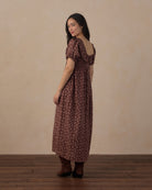 A woman wearing a brown and beige floral midi dress with puff sleeves and a low back, standing in profile against a neutral beige wall and dark wood floor.