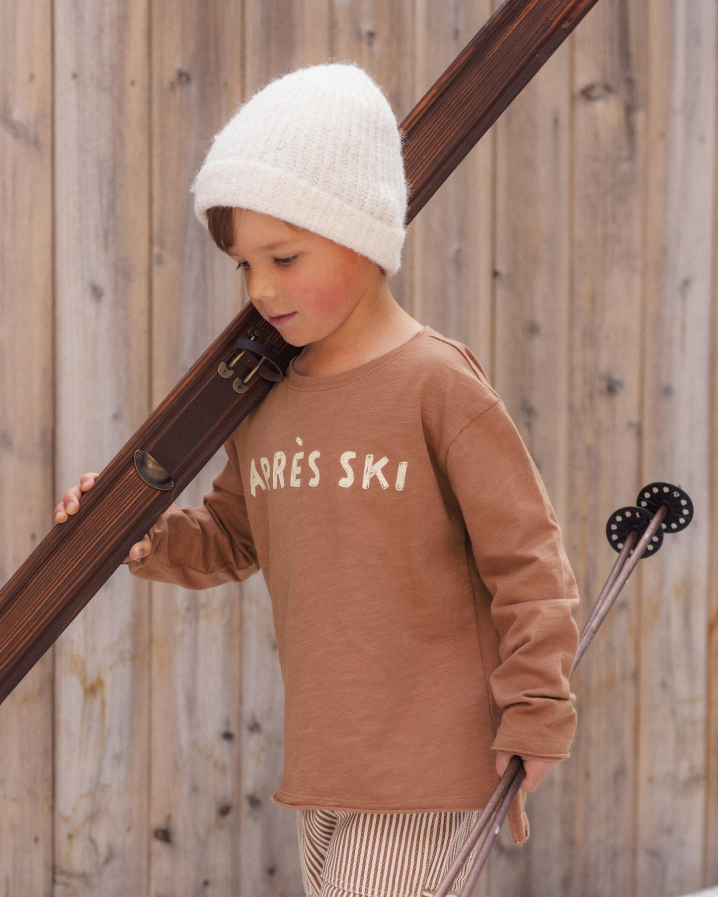A young child wearing a brown long-sleeve shirt with 'ARRÈS SKI' printed on the chest, a white knit beanie, and striped pants, holding ski poles against a wooden fence background.