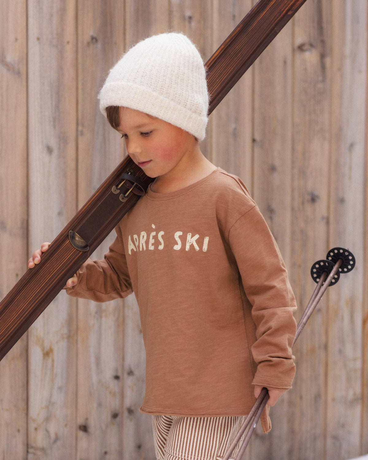 A young child wearing a brown long-sleeve shirt with 'ARRÈS SKI' printed on the chest, a white knit beanie, and striped pants, holding ski poles against a wooden fence background.