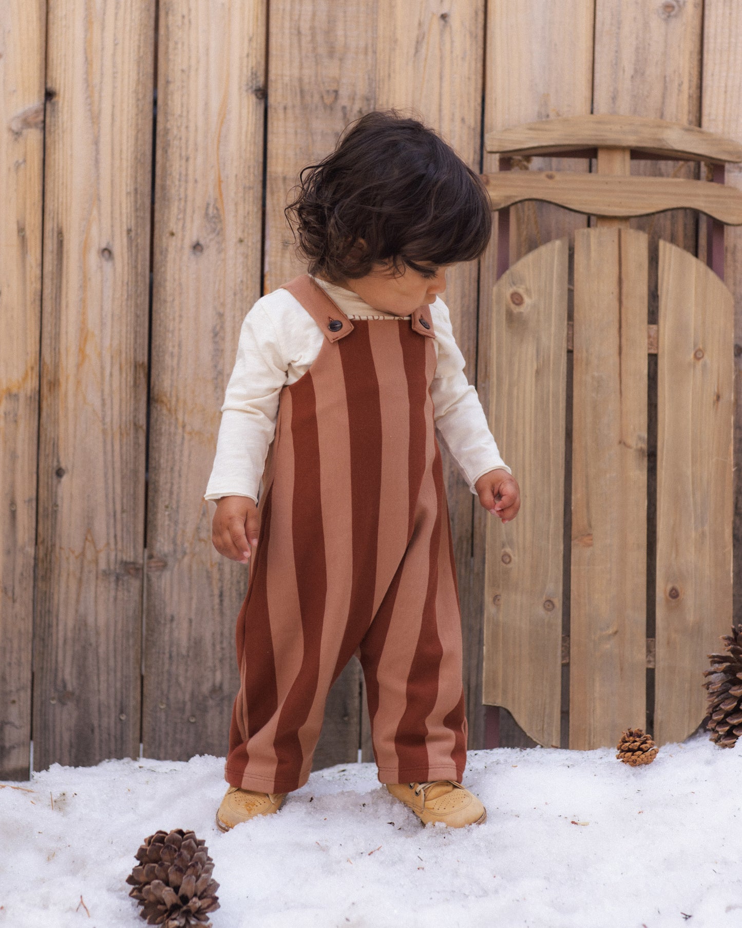 A toddler standing outdoors in the snow in front of a wooden fence, wearing brown and dusty rose vertical striped overalls over a cream long-sleeve shirt.