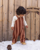 A toddler standing outdoors in the snow in front of a wooden fence, wearing brown and dusty rose vertical striped overalls over a cream long-sleeve shirt.