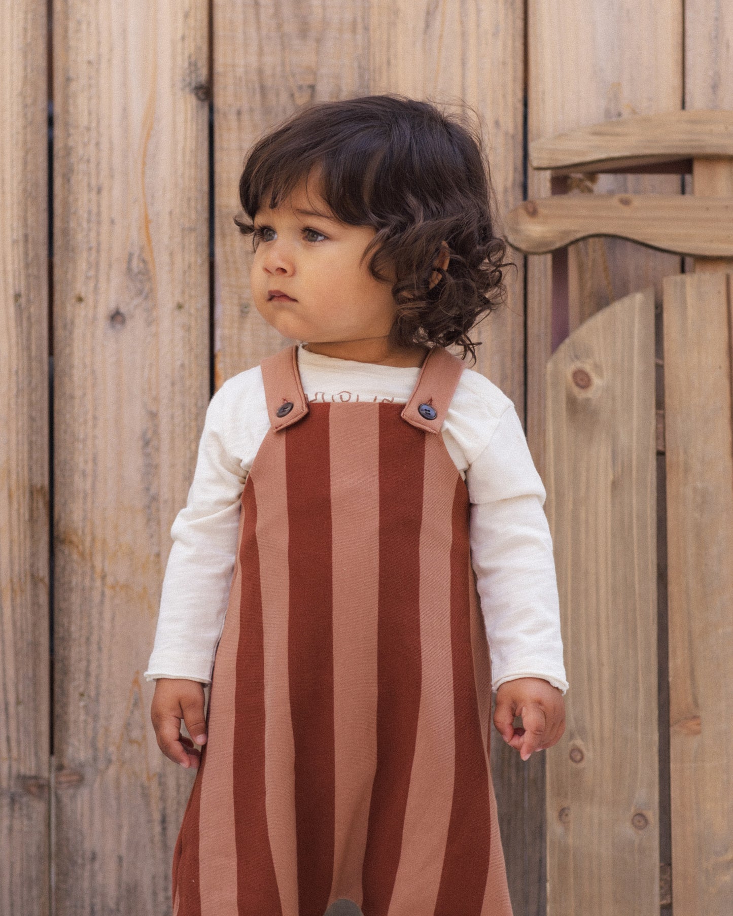 A toddler with dark curly hair stands in front of a rustic wooden fence, wearing a brown and tan vertically striped pinafore overalls outfit over a white long-sleeve shirt.