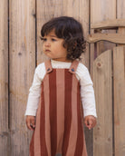 A toddler with dark curly hair stands in front of a rustic wooden fence, wearing a brown and tan vertically striped pinafore overalls outfit over a white long-sleeve shirt.