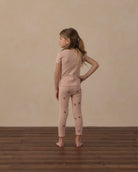 A young girl stands with her back to the camera wearing a light pink short-sleeve knit top and matching leggings with small brown star prints, set against a textured beige wall and dark wood floor.
