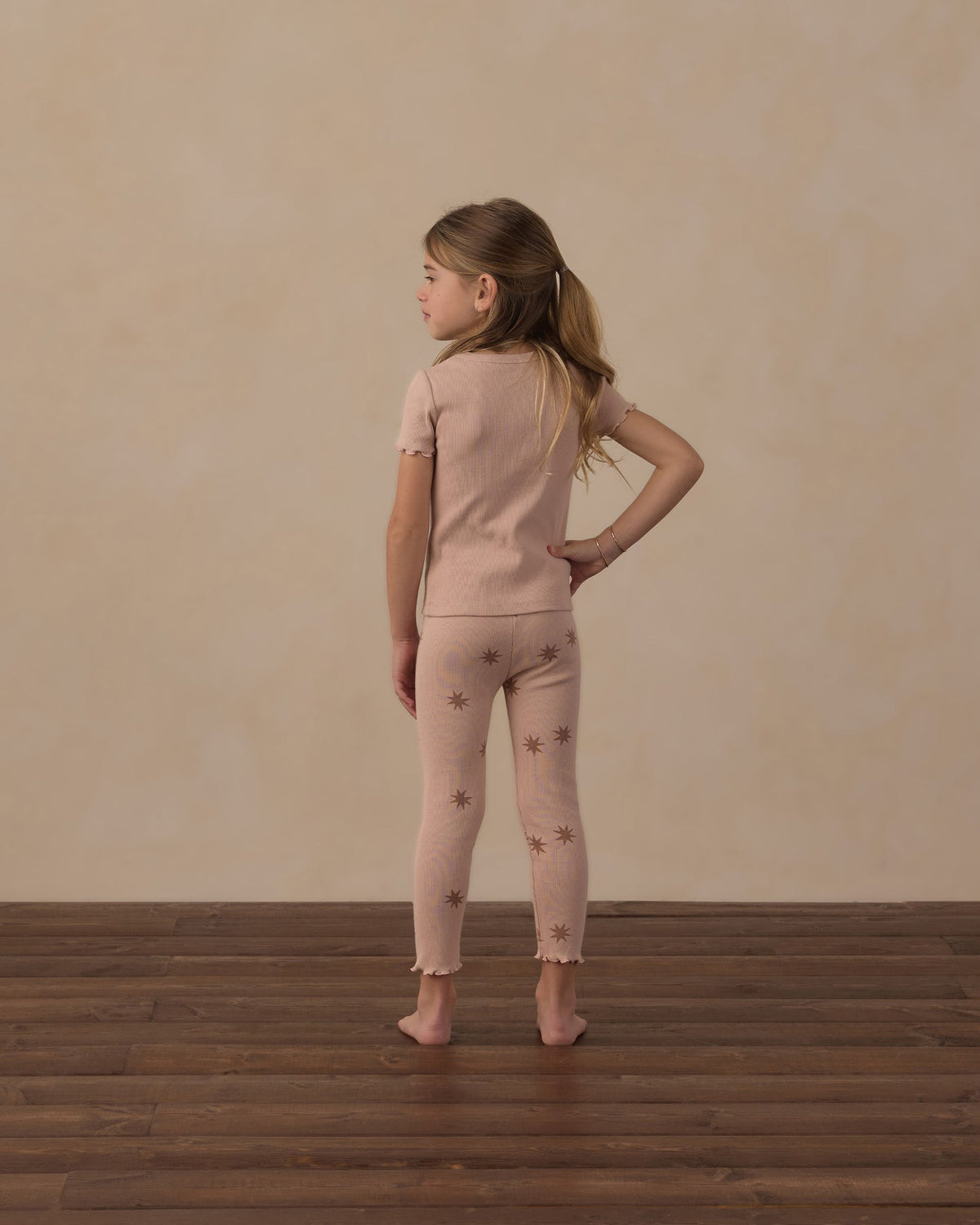A young girl stands with her back to the camera wearing a light pink short-sleeve knit top and matching leggings with small brown star prints, set against a textured beige wall and dark wood floor.