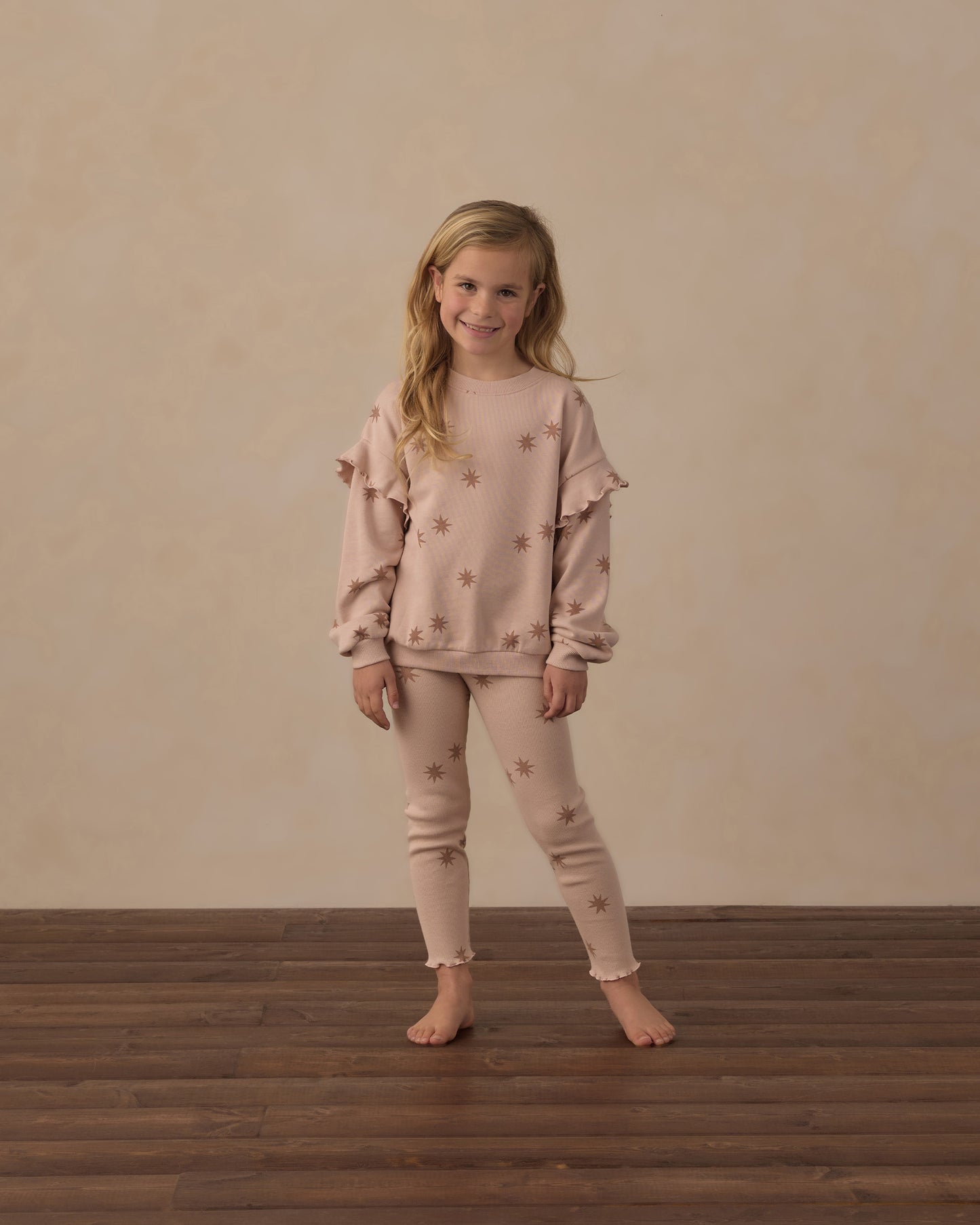 A young girl stands barefoot on a dark wood floor against a textured beige wall, wearing a matching dusty rose pajama set with star details.