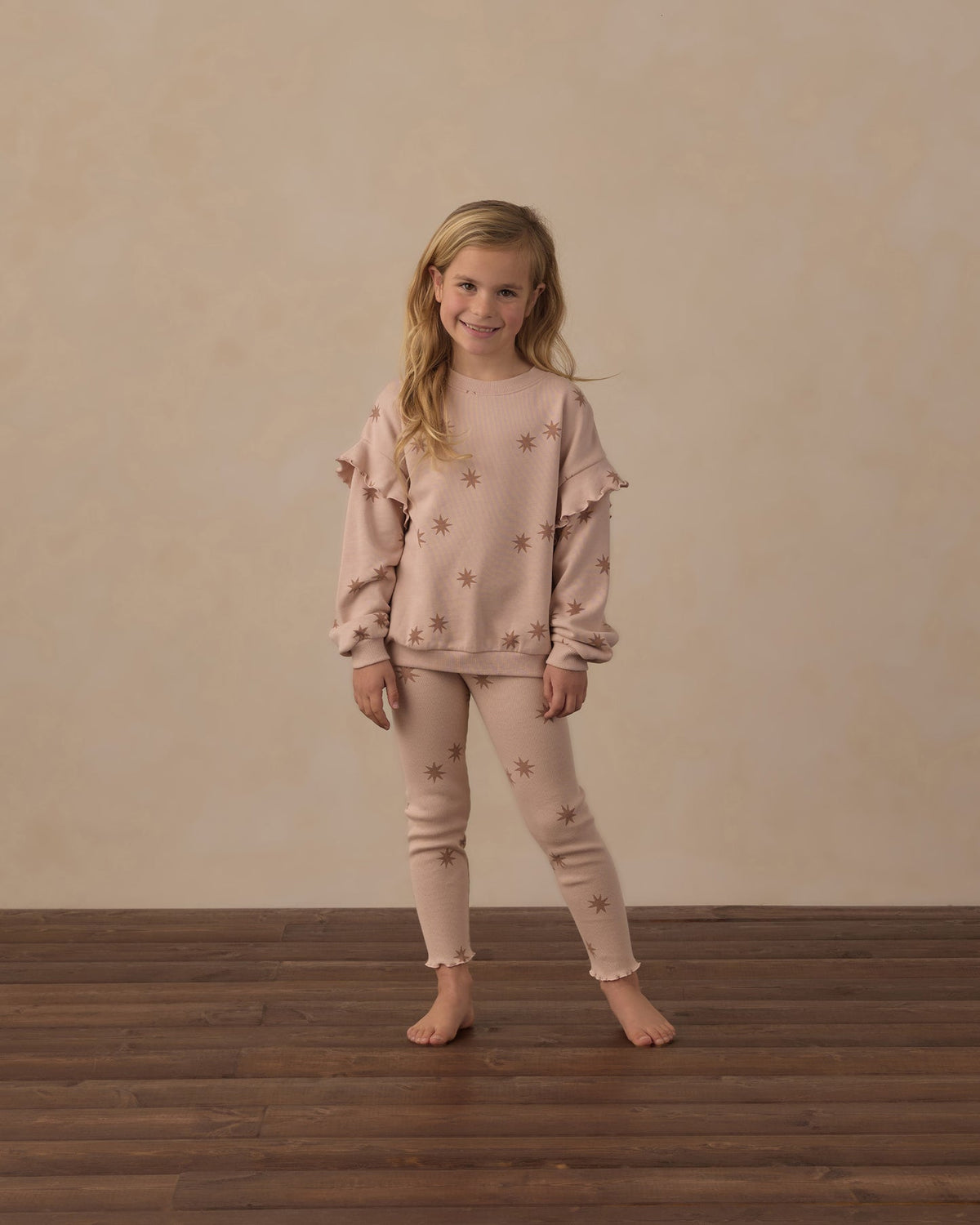 A young girl stands barefoot on a dark wood floor against a textured beige wall, wearing a matching dusty rose pajama set with star details.