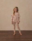 A young girl stands barefoot on a dark wood floor against a textured beige wall, wearing a matching dusty rose pajama set with star details.