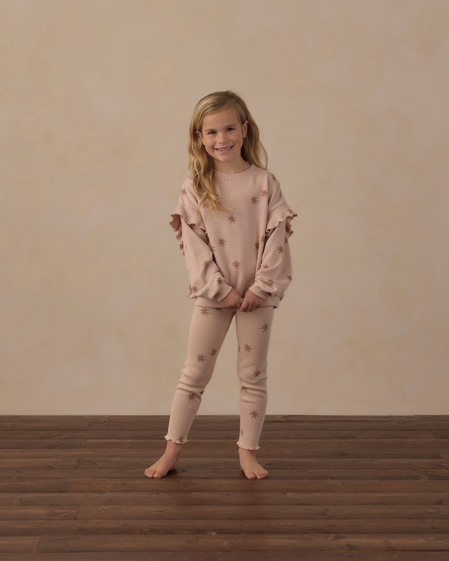 A young girl stands barefoot on a dark wood floor against a textured beige wall, wearing a matching dusty pink pajama set with star prints. The set includes a long-sleeve top with ruffled shoulders and matching leggings.