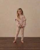 A young girl stands barefoot on a dark wood floor against a textured beige wall, wearing a matching dusty pink pajama set with star prints. The set includes a long-sleeve top with ruffled shoulders and matching leggings.