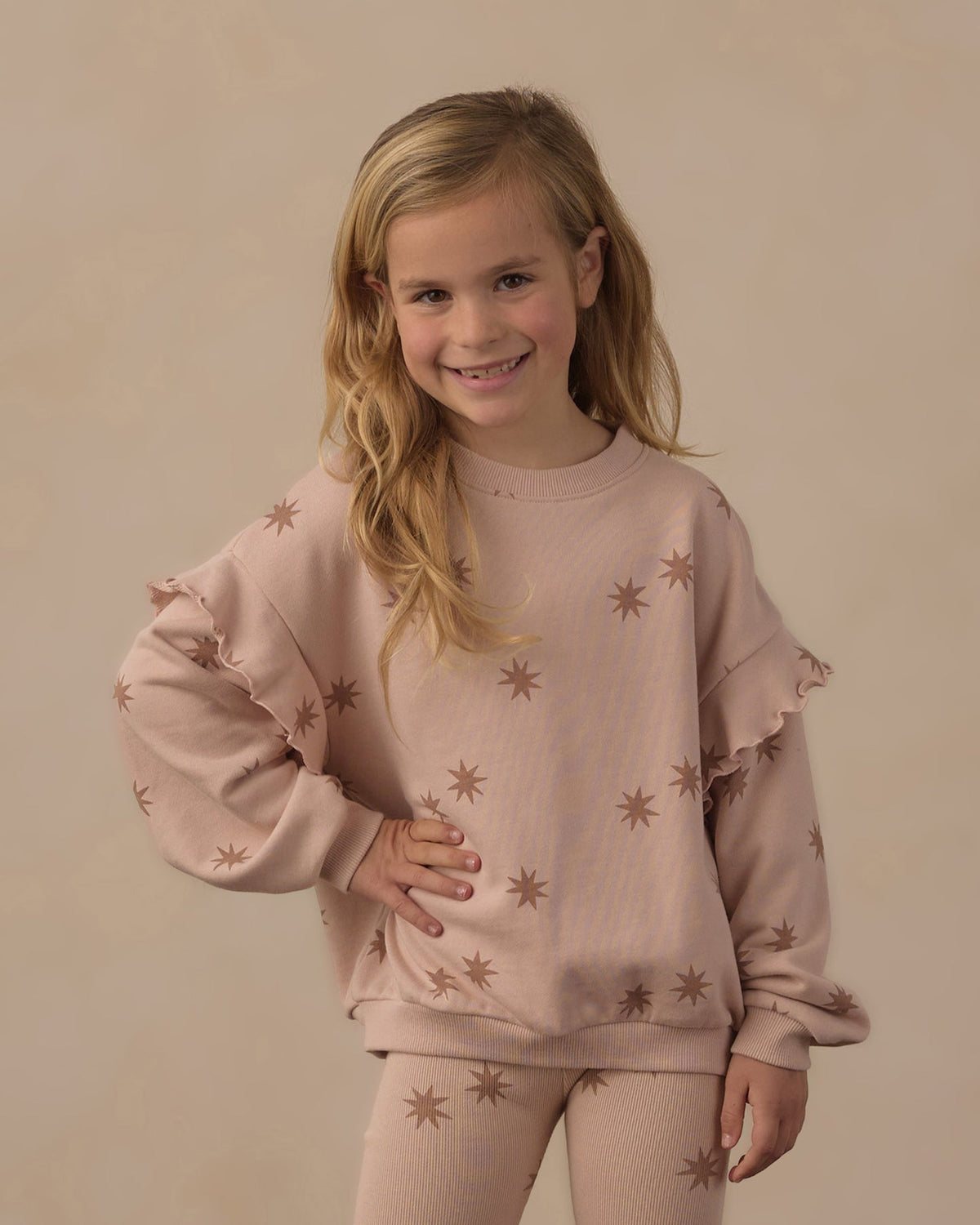 A young girl with blonde hair smiling, wearing a dusty pink long-sleeve sweatshirt with brown starburst patterns and matching ruffled sleeves, paired with matching leggings, against a solid beige background.