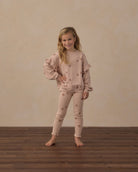 A young girl stands barefoot on a dark wood floor against a textured beige wall, wearing a matching light pink pajama set with star details.