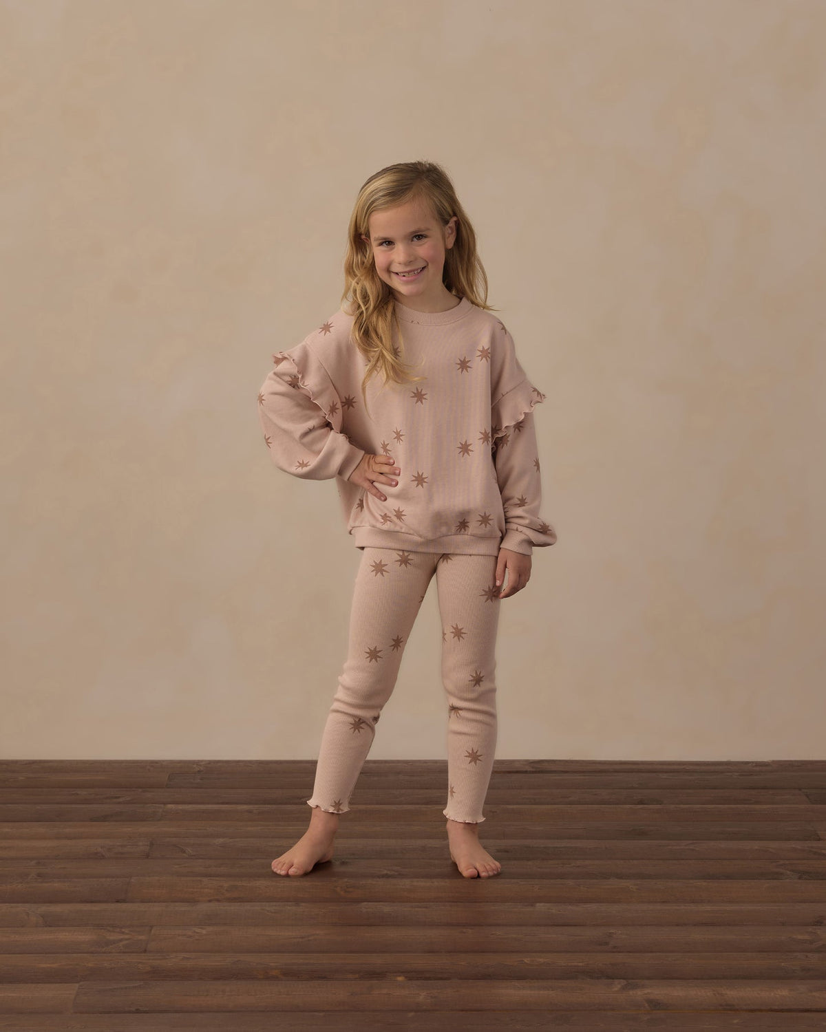 A young girl stands barefoot on a dark wood floor against a textured beige wall, wearing a matching light pink pajama set with star details.