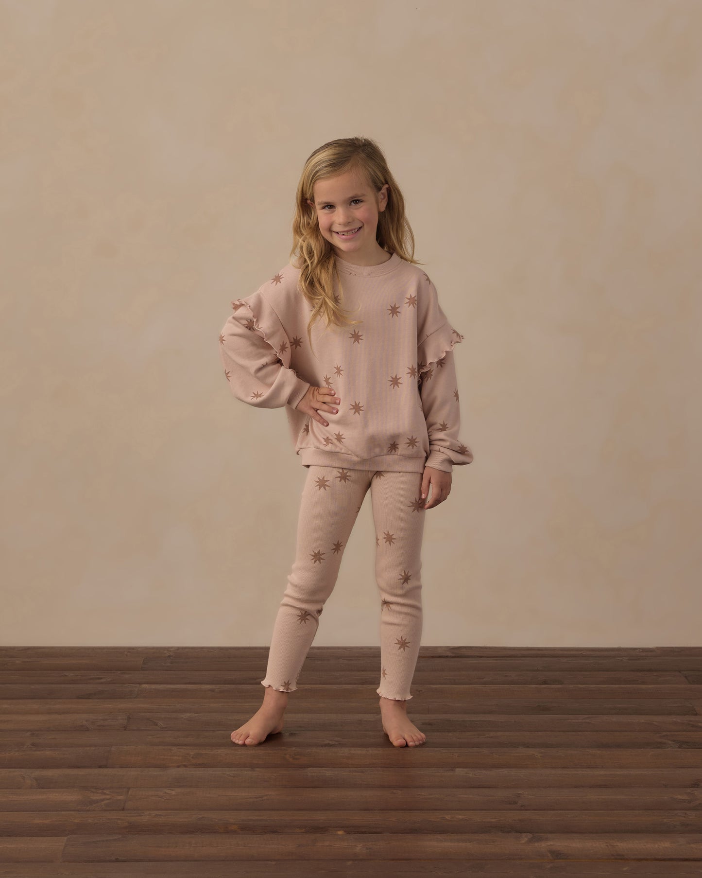 A young girl stands barefoot on a dark wood floor against a textured beige wall, wearing a matching light pink pajama set with star details.