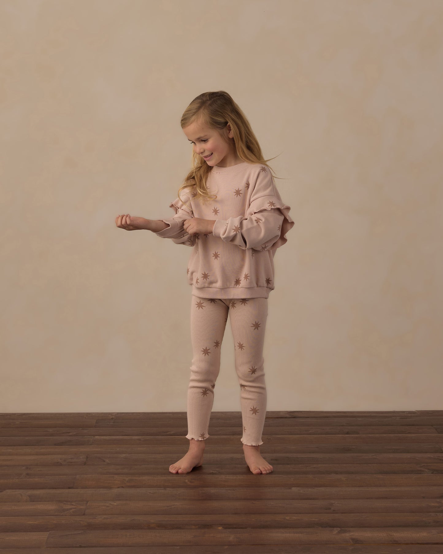 A young girl stands barefoot on a dark wood floor against a textured beige wall, wearing a matching light pink pajama set with small embroidered star details. She is adjusting the cuffs of her long-sleeve top.