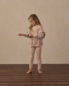 A young girl stands barefoot on a dark wood floor against a textured beige wall, wearing a matching light pink pajama set with small embroidered star details. She is adjusting the cuffs of her long-sleeve top.