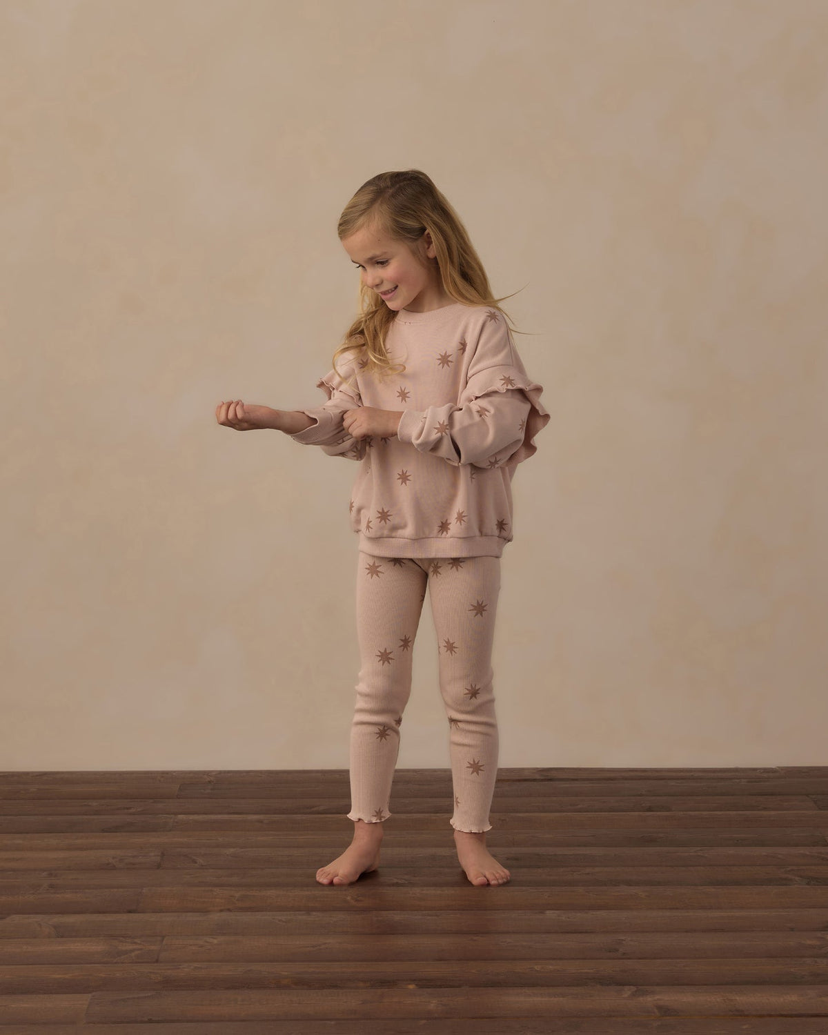A young girl stands barefoot on a dark wood floor against a textured beige wall, wearing a matching light pink pajama set with small embroidered star details. She is adjusting the cuffs of her long-sleeve top.
