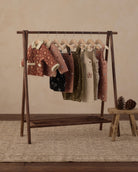 A clothing rack displaying multiple pairs of overall in different colors and patterns, including pink, olive green, beige, and black, hanging on wooden hangers against a warm beige background.