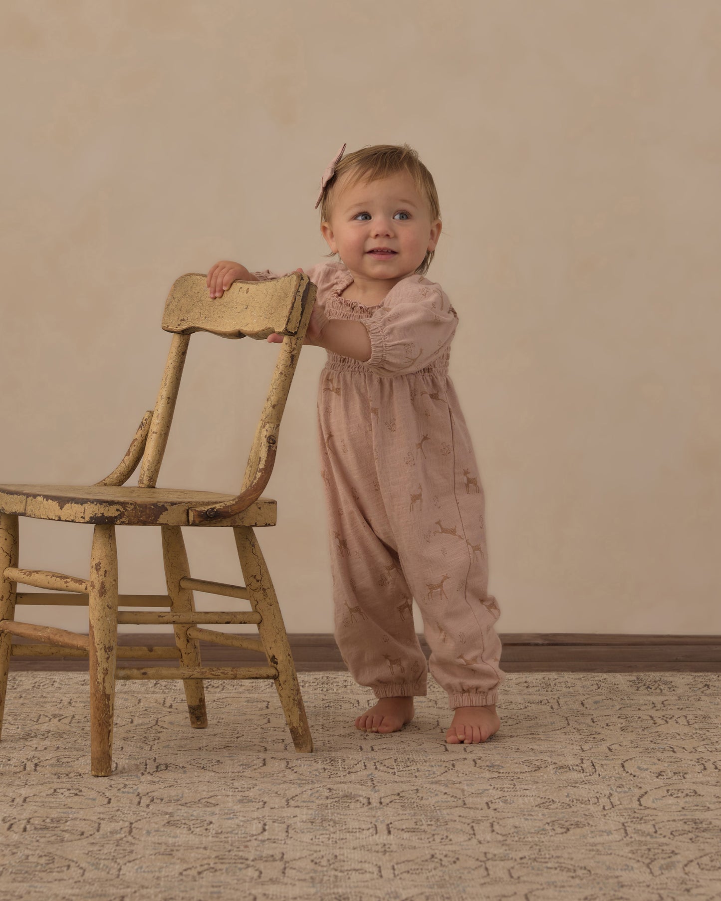 A baby standing on a patterned rug next to a rustic wooden chair, wearing a dusty pink long-sleeve jumpsuit with a subtle star pattern.